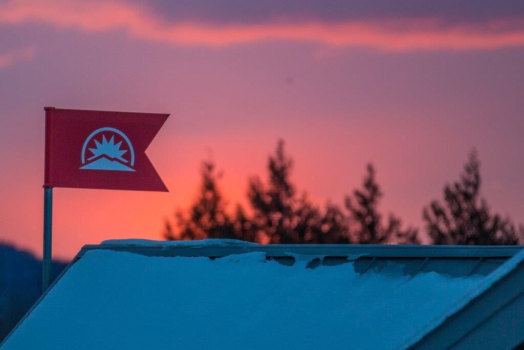 Eine Fahne mit Wappen, das eine Berg- und Sonnensymbol zeigt, auf einem Mast vor einem Sonnenuntergangshimmel.