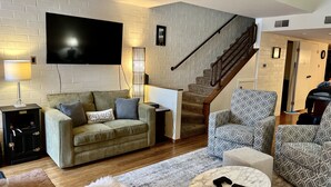 Ein Wohnzimmer mit einer Couch, einem gemusterten Sessel und einem Couchtisch. Ein Fernseher ist an der Wand befestigt, und im Hintergrund ist eine Treppe zu sehen.