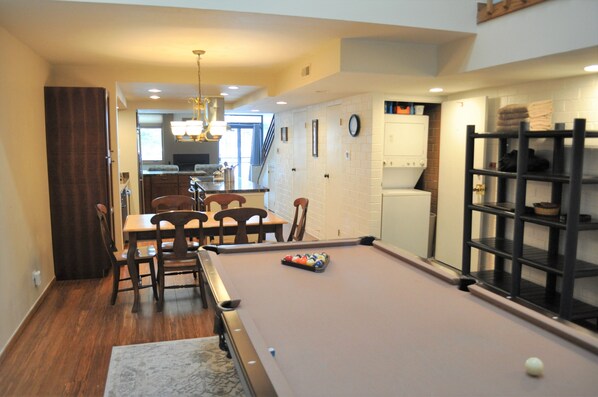 Une table de billard dans une pièce qui comprend un coin repas, une cuisine à l’arrière-plan et un escalier menant à un autre niveau.