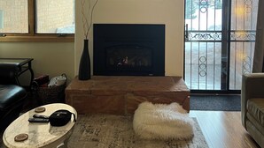 Ein Wohnzimmer mit Kamin, einem steinernen Kaminumbau, einem Marmortisch, einem flauschigen weißen Pouf und Blick durch eine Glastür auf einen Balkon.