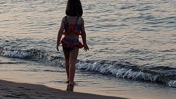 Une scène de plage où un enfant marche vers l’eau, un phare se dressant au loin et les vagues venant doucement lécher le rivage.