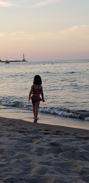 Une scène de plage où un enfant marche vers l’eau, un phare se dressant au loin et les vagues venant doucement lécher le rivage.