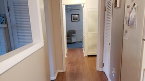 Un couloir étroit avec un plancher en bois, des murs blancs et une porte donnant sur une pièce équipée d’un ventilateur de plafond et d’un canapé.