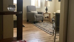 Ein Wohnzimmer mit Holzboden, einem grauen Sessel, einem kleinen Tisch mit einer Lampe, einem gemusterten Teppich und einer karierten Tischdecke.