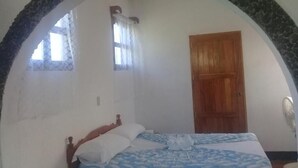 Un dormitorio con un arco de piedra, una puerta de madera, una cama con colcha de rayas azules y blancas, y una ventana con cortinas.