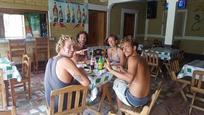 Un grupo de personas cenando en un restaurante con sillas y mesas de madera, suelo de baldosas y una pared con un anuncio de bebidas.