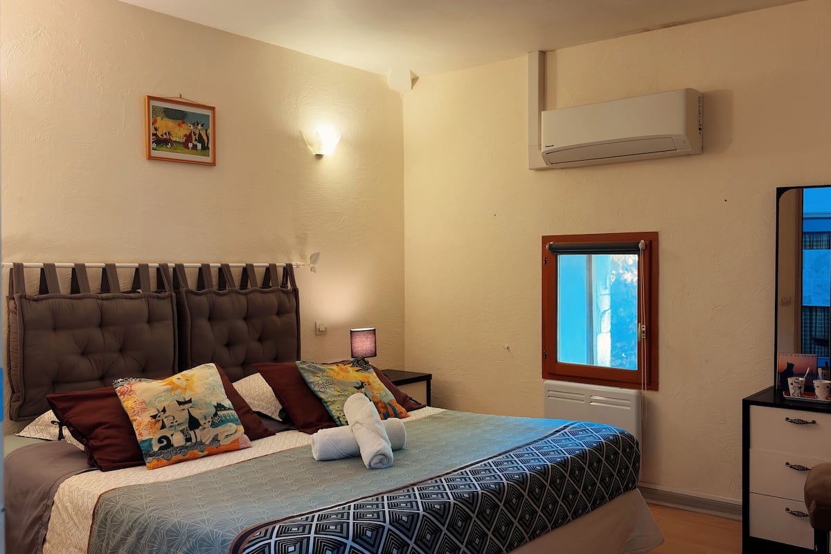 A bedroom with a bed, a nightstand, a dresser, an air conditioning unit, a window, and a wall-mounted light fixture.