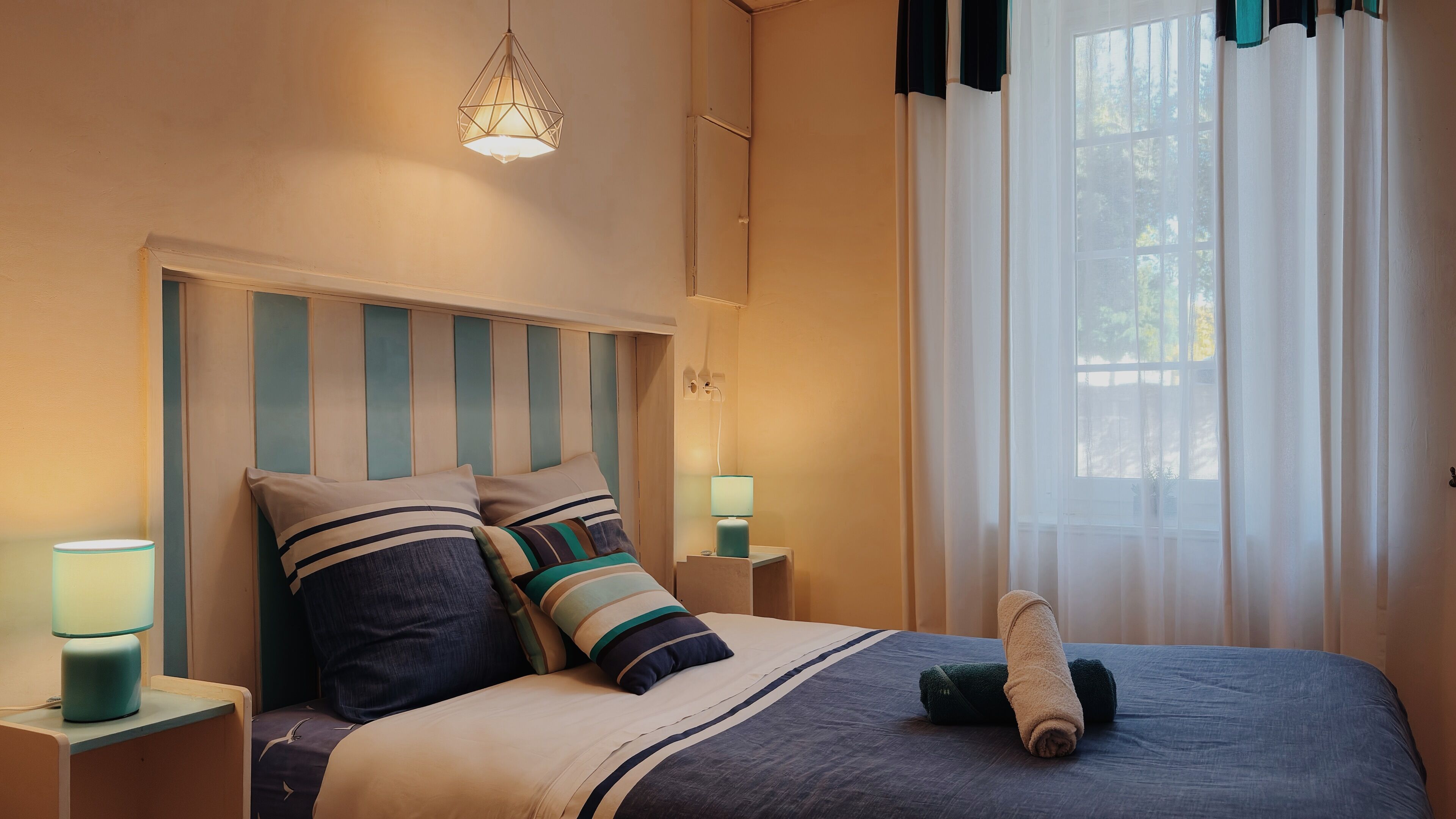 Una cama bien hecha con ropa de cama a rayas, una lámpara de noche y una ventana con cortinas translúcidas.