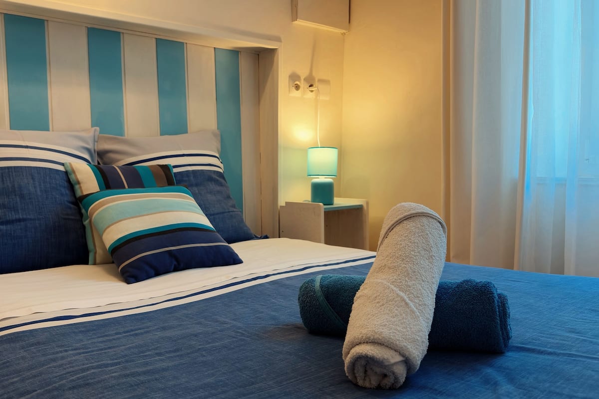 A neatly made bed with striped pillows and a rolled towel, a bedside lamp, and a view of a window with curtains.
