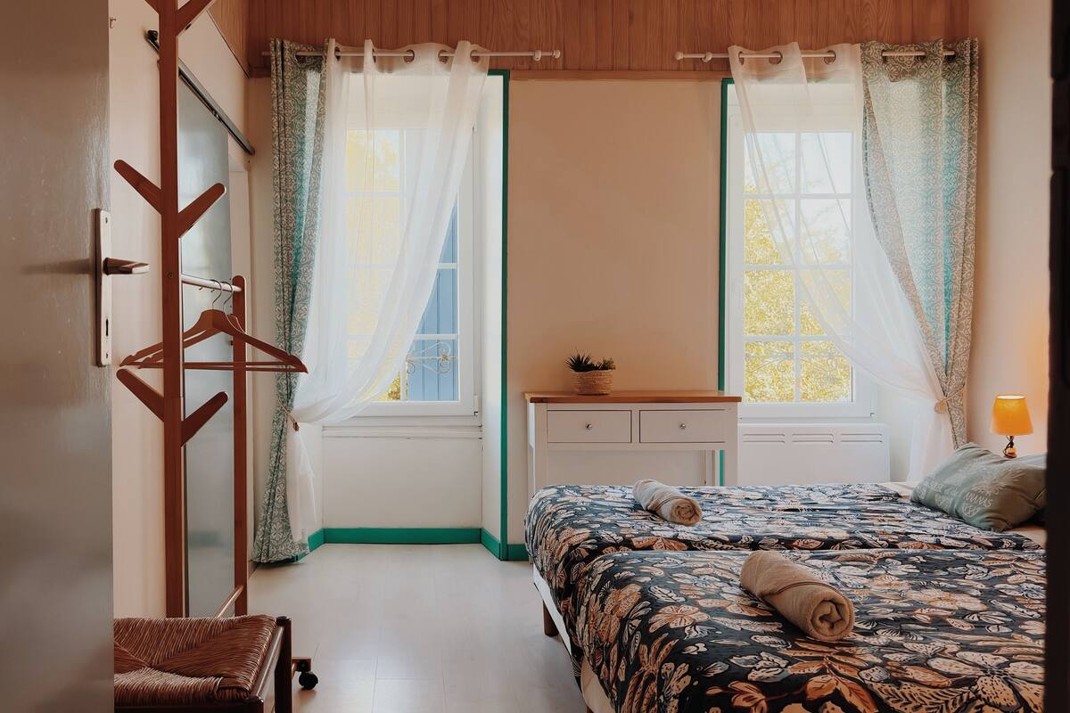 A bedroom with a bed, a bedside table, a hanging coat rack, and a window with curtains.