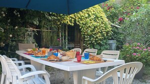 Un espace de restauration en plein air avec une table dressée pour le petit-déjeuner, entourée de chaises en plastique blanches et protégée par un parasol bleu.