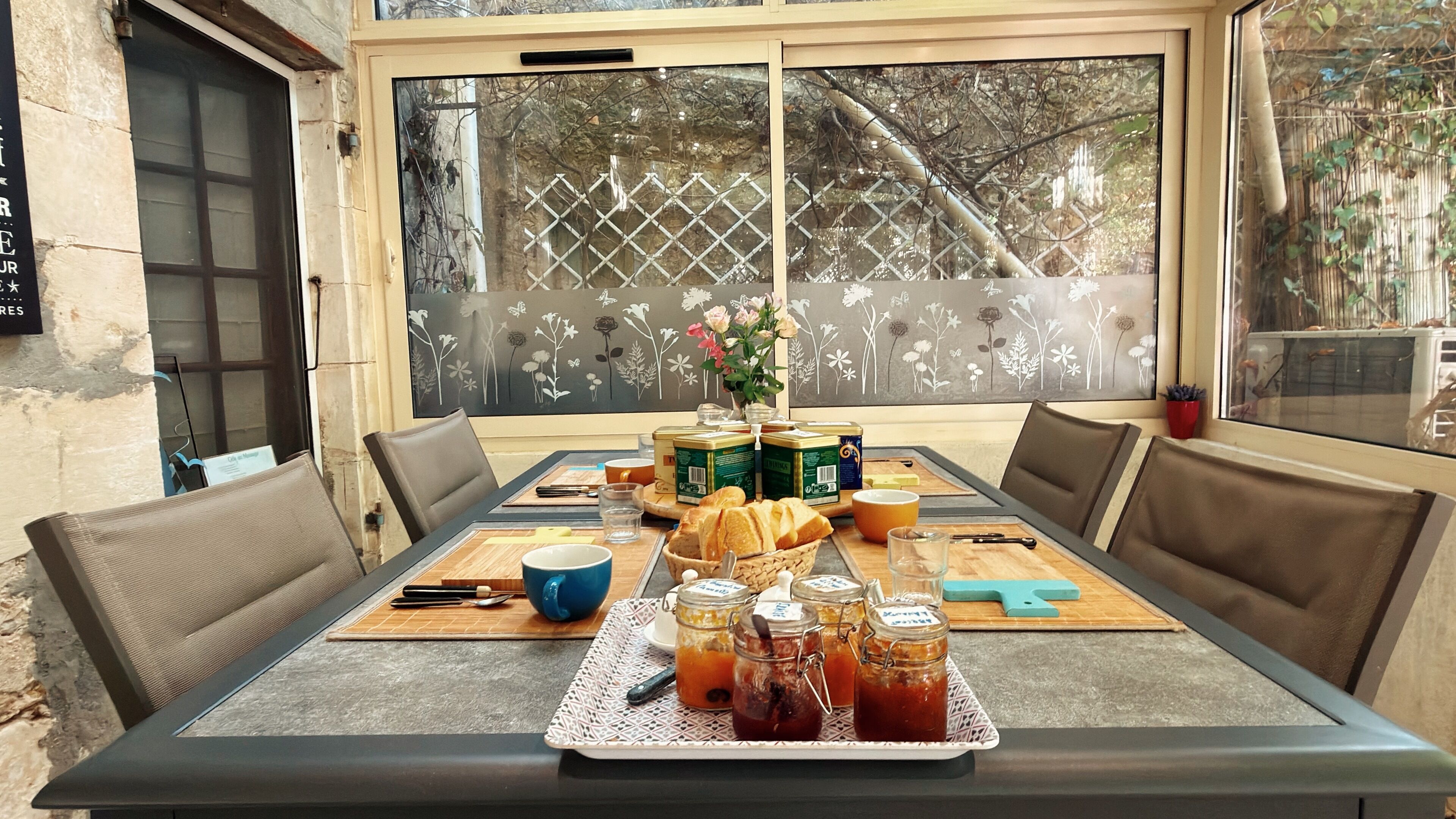 Une table à manger dressée avec du pain, des pots de confiture et un vase de fleurs.
