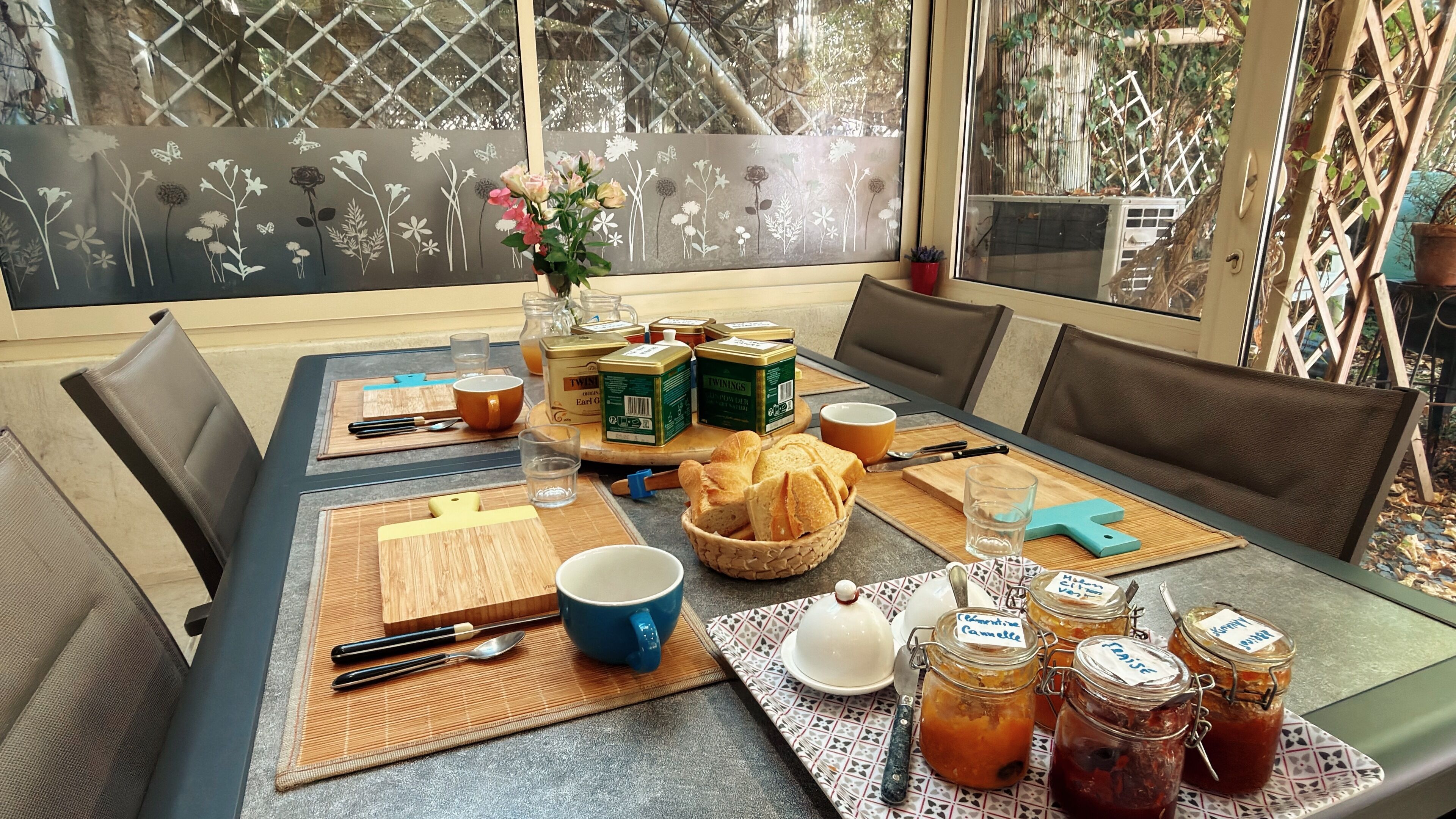 Une table à manger dressée avec de la nourriture et des boissons, entourée de chaises, dans une pièce donnant sur un paysage grâce à une fenêtre à treillis.