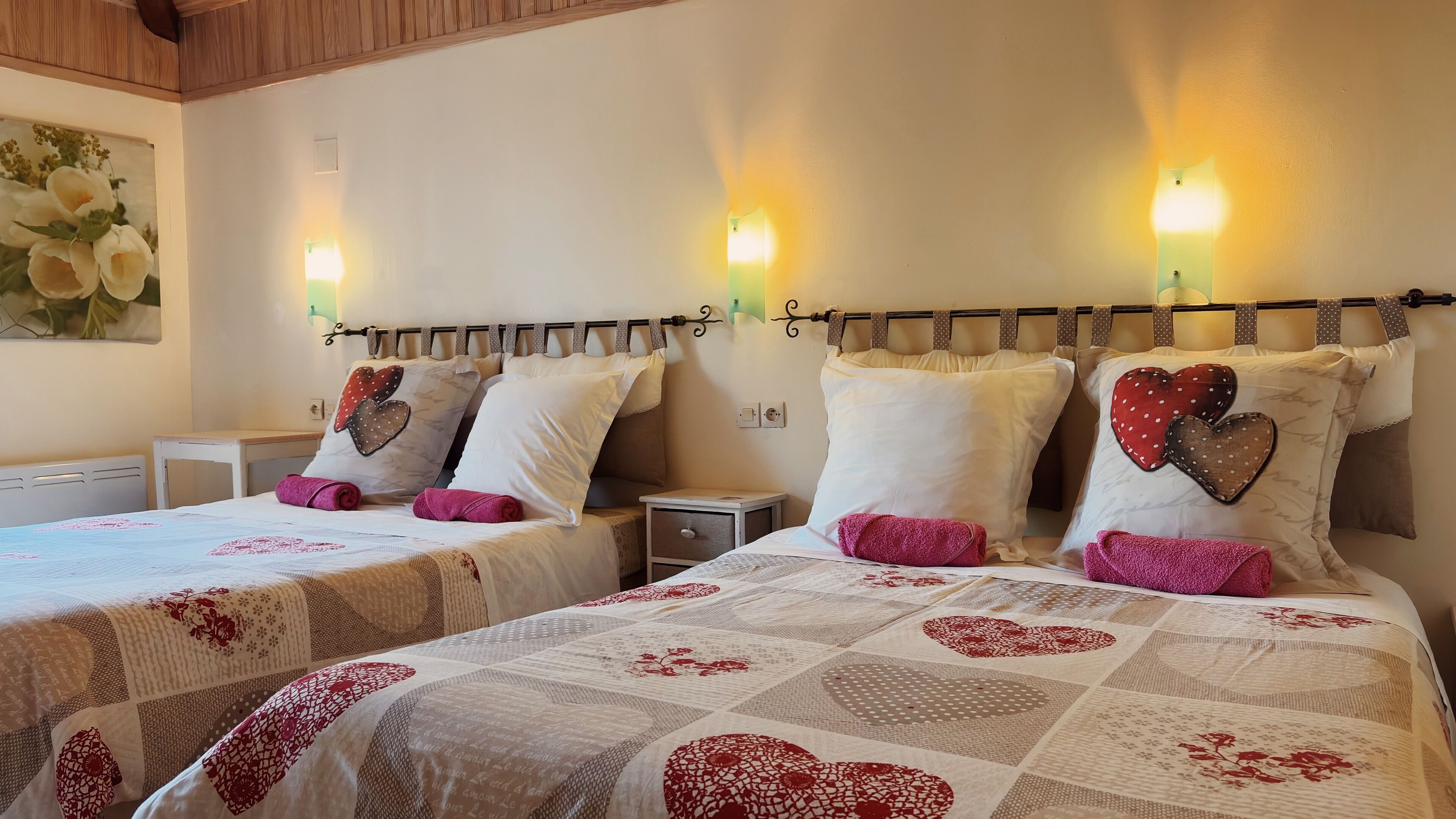 A hotel room with two beds, each with a quilted bedspread featuring heart patterns. There are wall-mounted light fixtures and a small bedside table with a drawer.