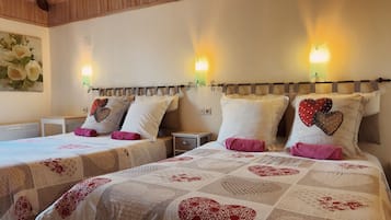 A hotel room with two beds, each with a quilted bedspread featuring heart patterns. There are wall-mounted light fixtures and a small bedside table with a drawer.