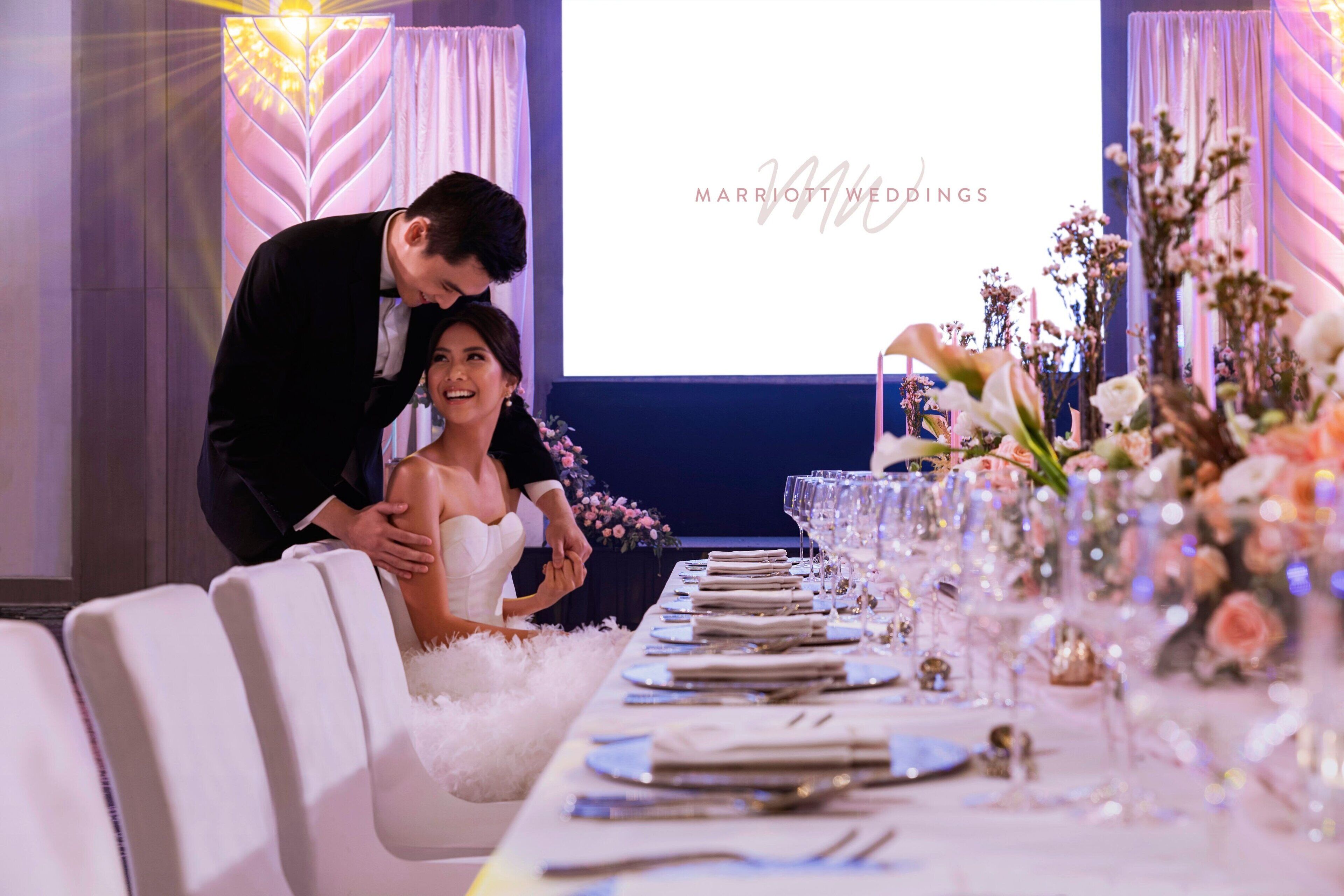 Una recepción de boda con una larga mesa dispuesta para los invitados, una pantalla que muestra 'MARRIOTT WEDDINGS' y adornos florales.