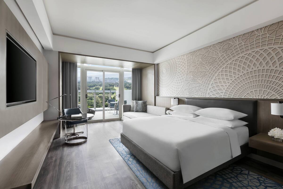 A modern hotel room with a large bed, a flat-screen TV, a desk with a chair, and a view of the city.