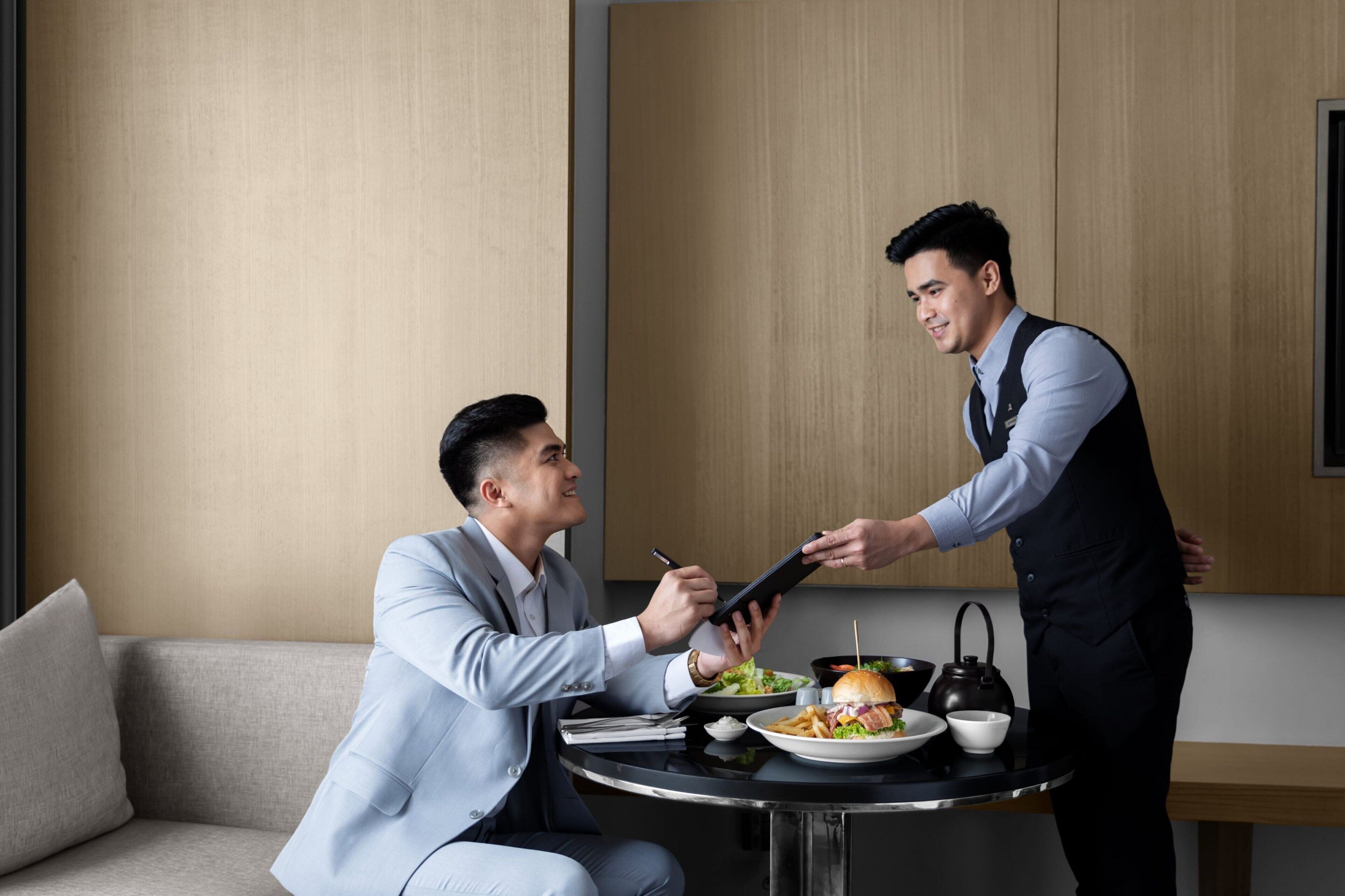 A man in a suit sitting at a round table with a burger and a drink, while another man in a vest stands beside him.