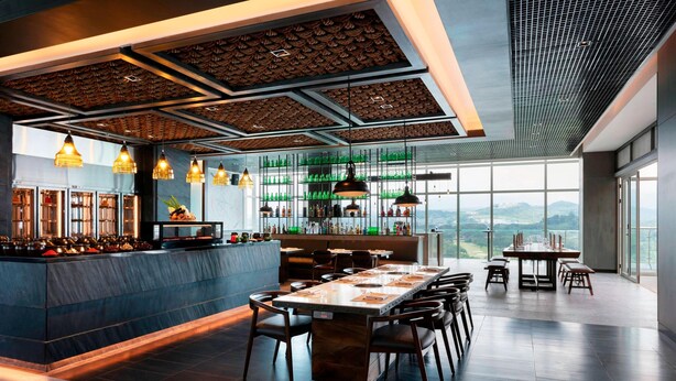 A modern restaurant with a long dining table, multiple chairs, a bar area with shelves of bottles, and large windows offering a view of the mountains. - Clark Marriott Hotel (Mabalacat City)