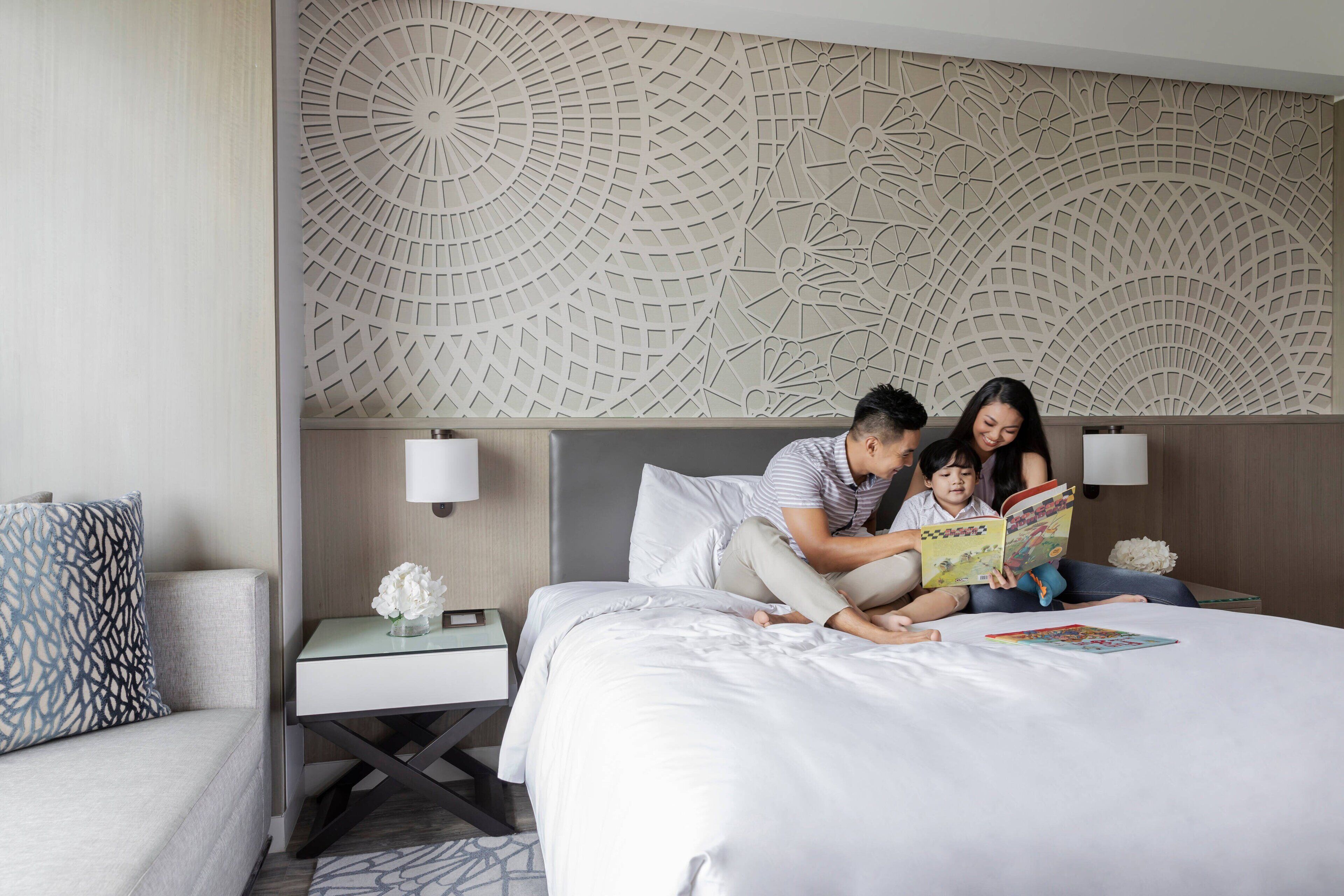 Habitación de hotel con cama, mesita de noche con lámpara, pared decorativa y sofá con cojín estampado.
