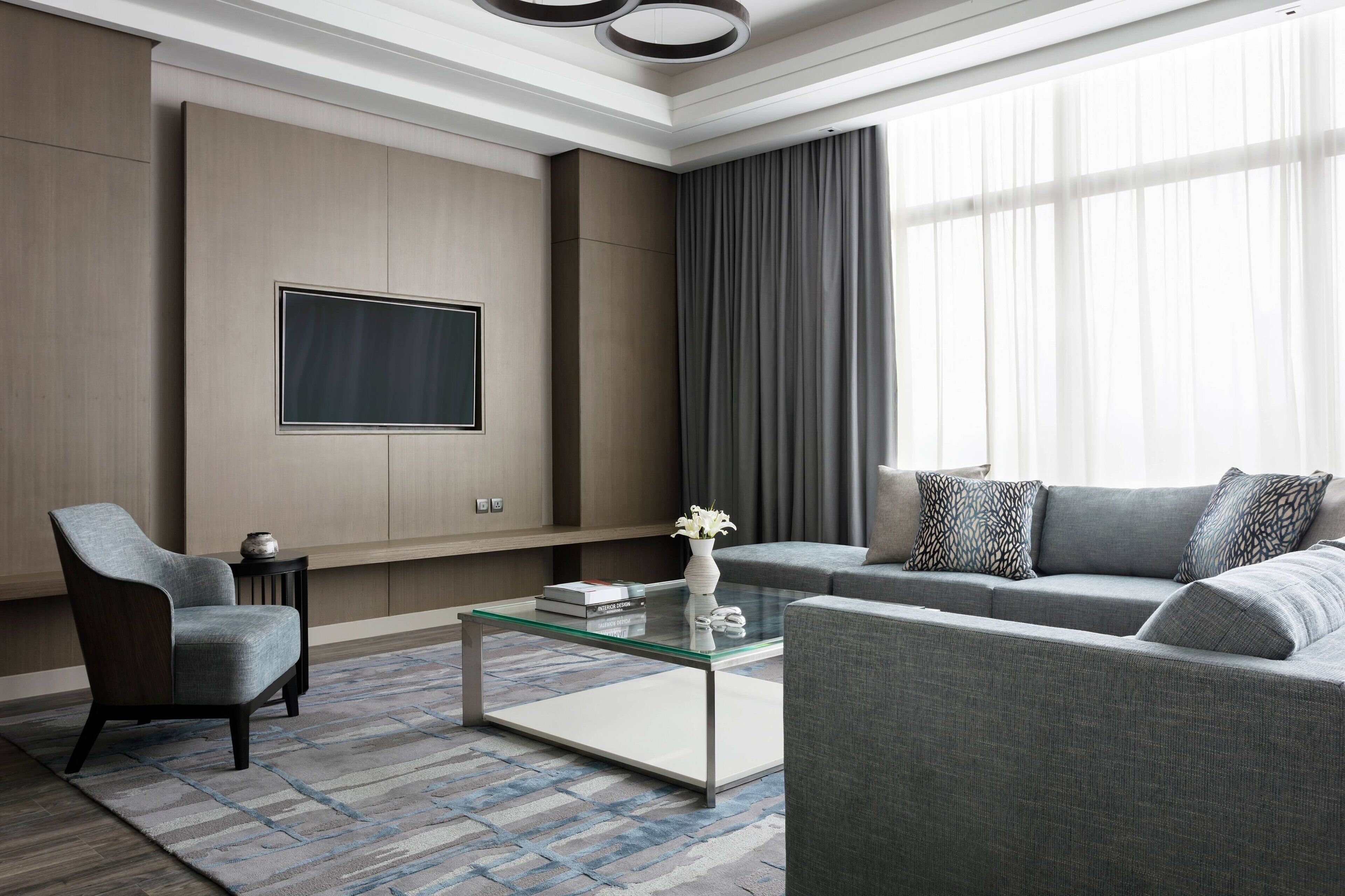 Una sala de estar moderna con una mesa de centro de vidrio, un sofá gris y un sillón, un televisor de pantalla plana montado en la pared y un ventanal con cortinas.