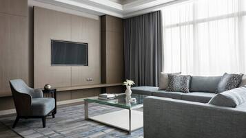 Una sala de estar moderna con una mesa de centro de vidrio, un sofá gris y un sillón, un televisor de pantalla plana montado en la pared y un ventanal con cortinas.