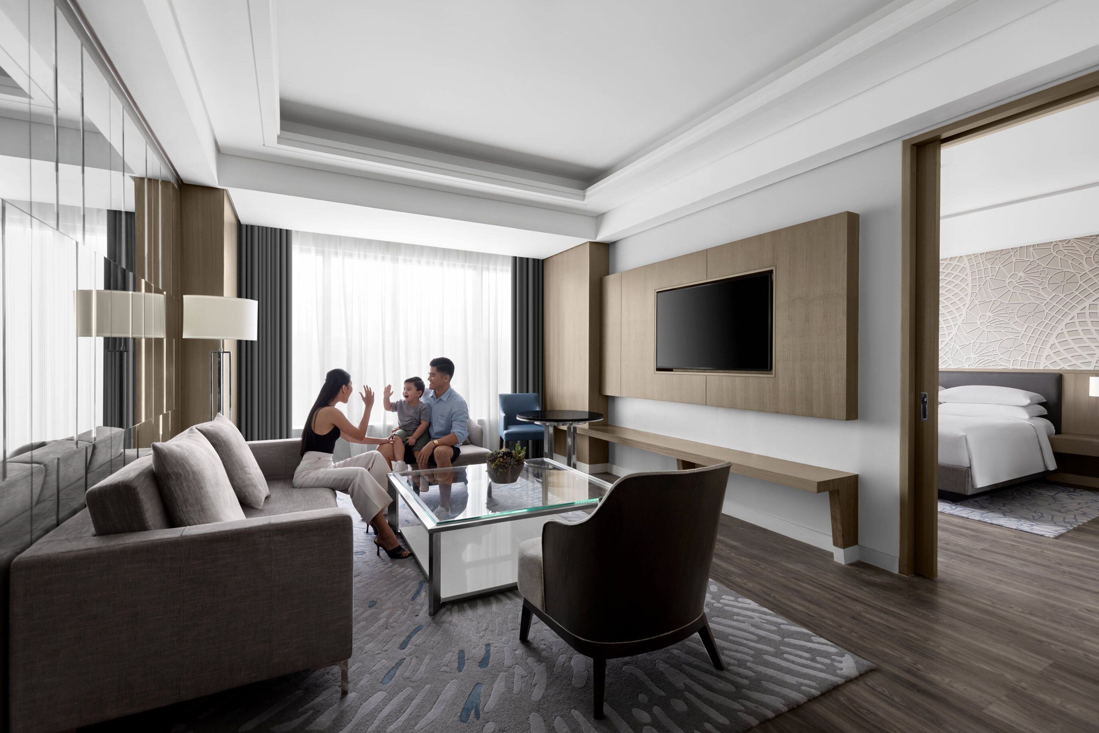 A modern hotel room with a glass coffee table, a sofa, a chair, a TV mounted on the wall, and a bed with a patterned rug.