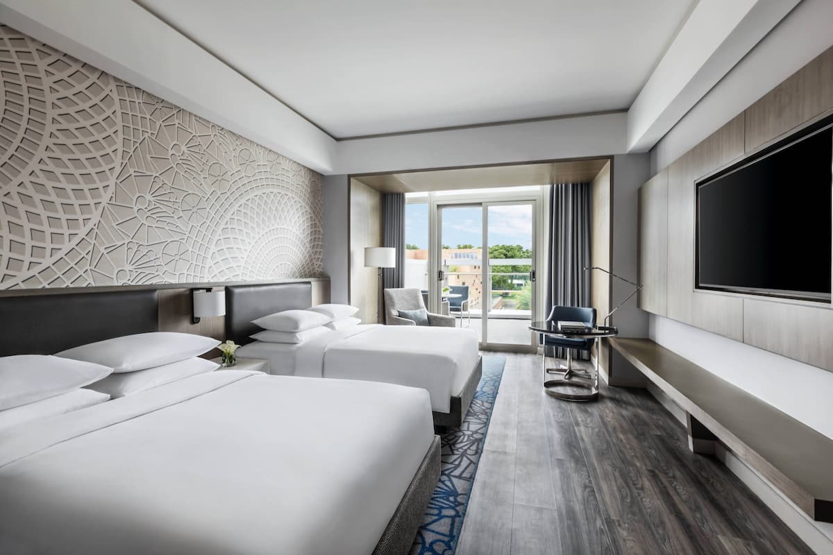 A hotel room with two beds, a large TV, a desk with a chair, and a view of the outdoors.