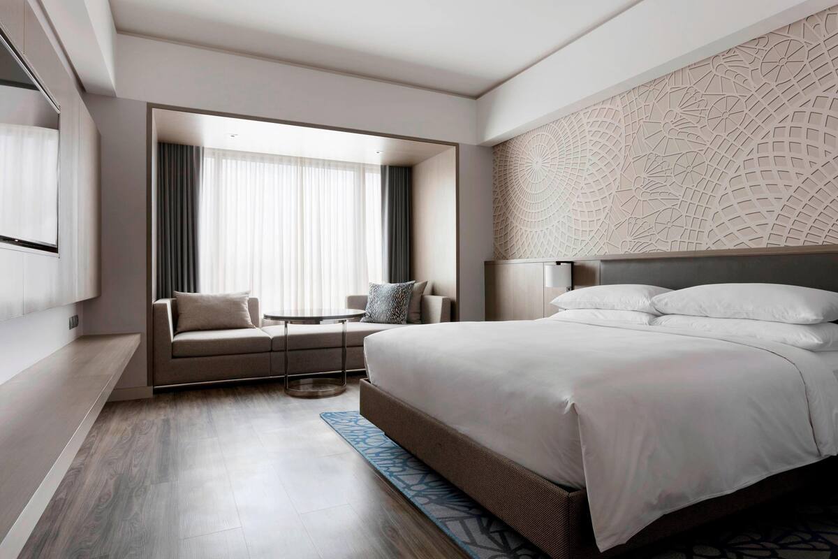 A modern hotel room with a large bed, a sofa, a small table, and a window with curtains.