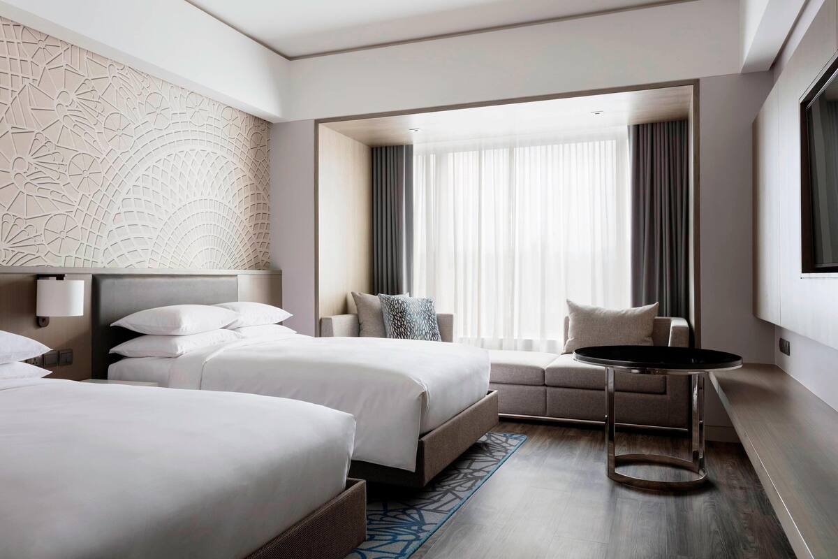 A modern hotel room with two beds, a sofa, a small round table, and a large window with curtains.