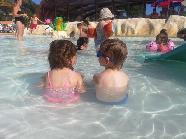 A pool area with clear water, a rock wall, and people swimming and playing. - Plaisirs Nature à La Pignade (La Tremblade)