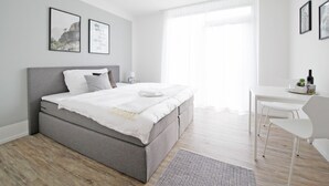 A modern bedroom with a grey bed, white bedding, a bedside table with a lamp, and a desk with chairs. - VARIAS Business Apartments (Elsau)