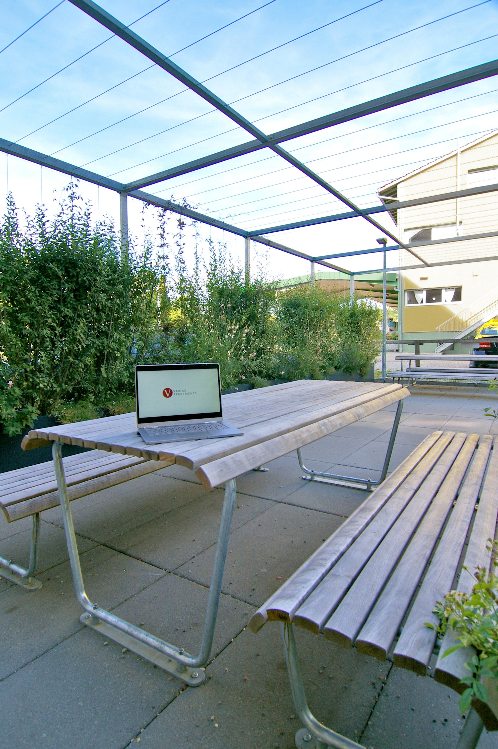 Un área de estar al aire libre con una mesa y bancos, una computadora portátil sobre la mesa y un edificio al fondo.