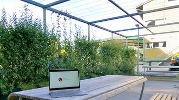 Un área de estar al aire libre con una mesa y bancos, una computadora portátil sobre la mesa y un edificio al fondo.