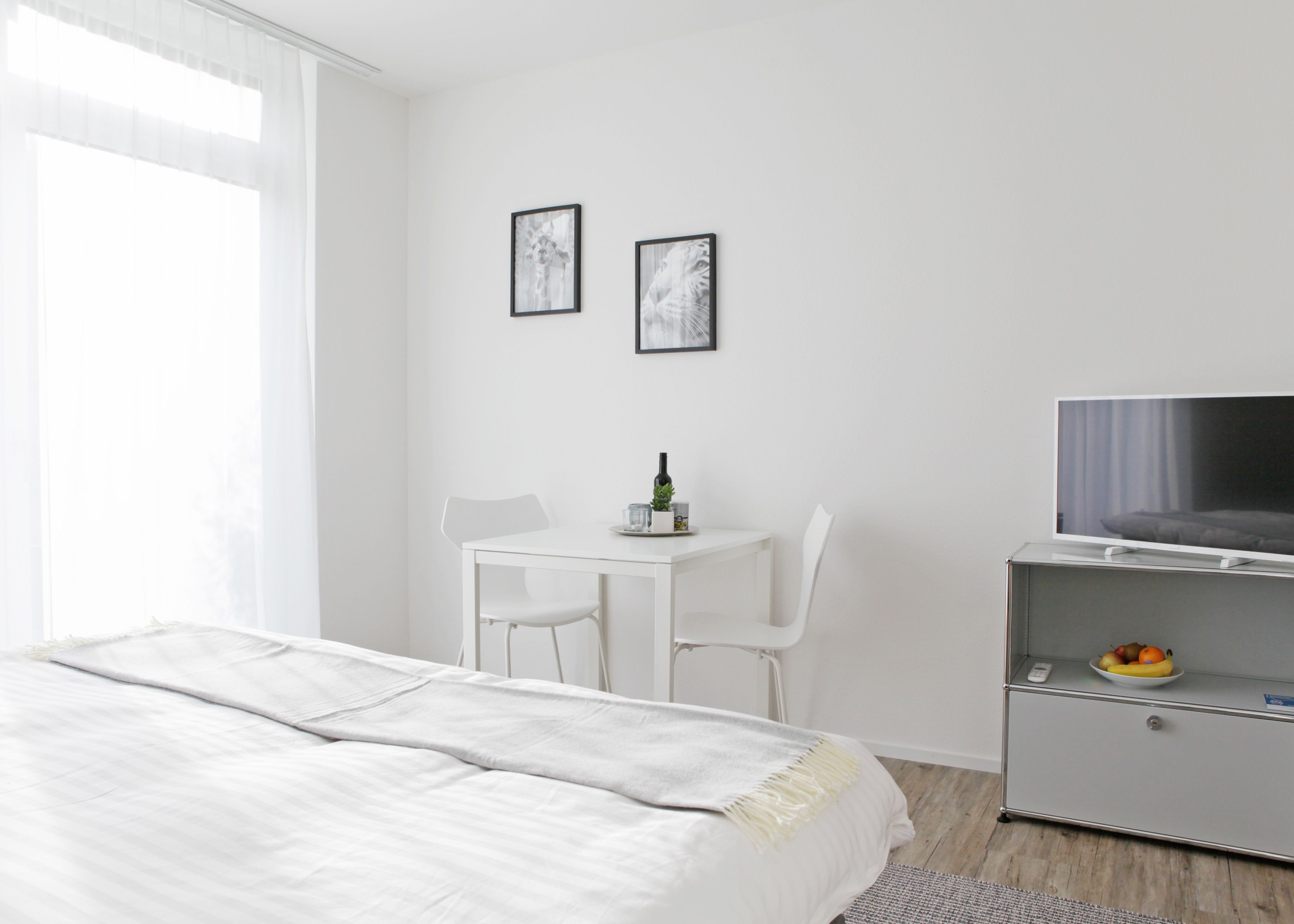 Ein schlicht eingerichtetes Hotelzimmer mit einem Bett, einem kleinen Tisch mit Pflanze, einem Fernseher auf einem Schrank und zwei gerahmten Bildern an der Wand.