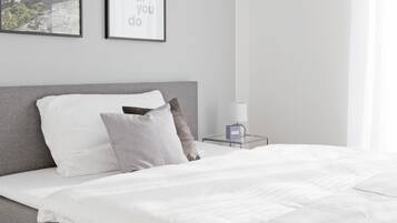 A neatly made bed with white linens and a grey headboard, a bedside table with a lamp and a decorative item, and two framed motivational quotes on the wall.