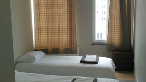 Una habitación de hotel con dos camas individuales, una ventana con cortinas, un radiador y una mesita pequeña.