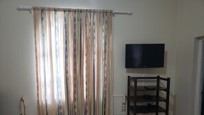 A room with a television mounted on the wall, a wooden chair, and a curtain with a geometric pattern. - The Barbican Haven (Kingston)