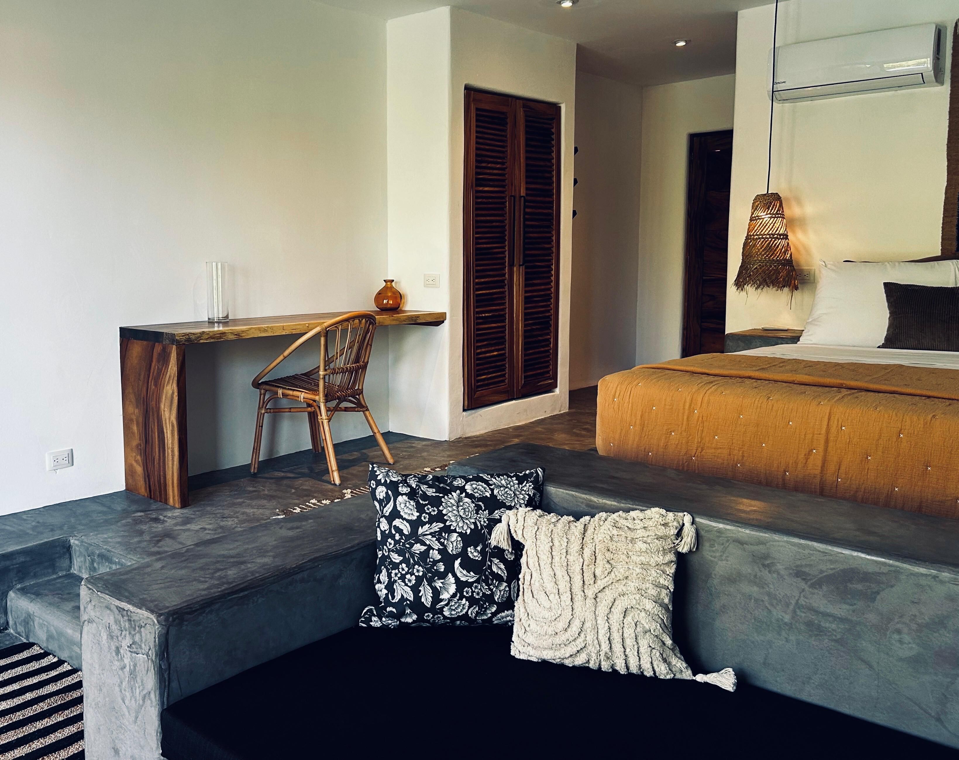 A bedroom with a bed, a desk, and a chair. There is a window with shutters and an air conditioning unit on the wall.