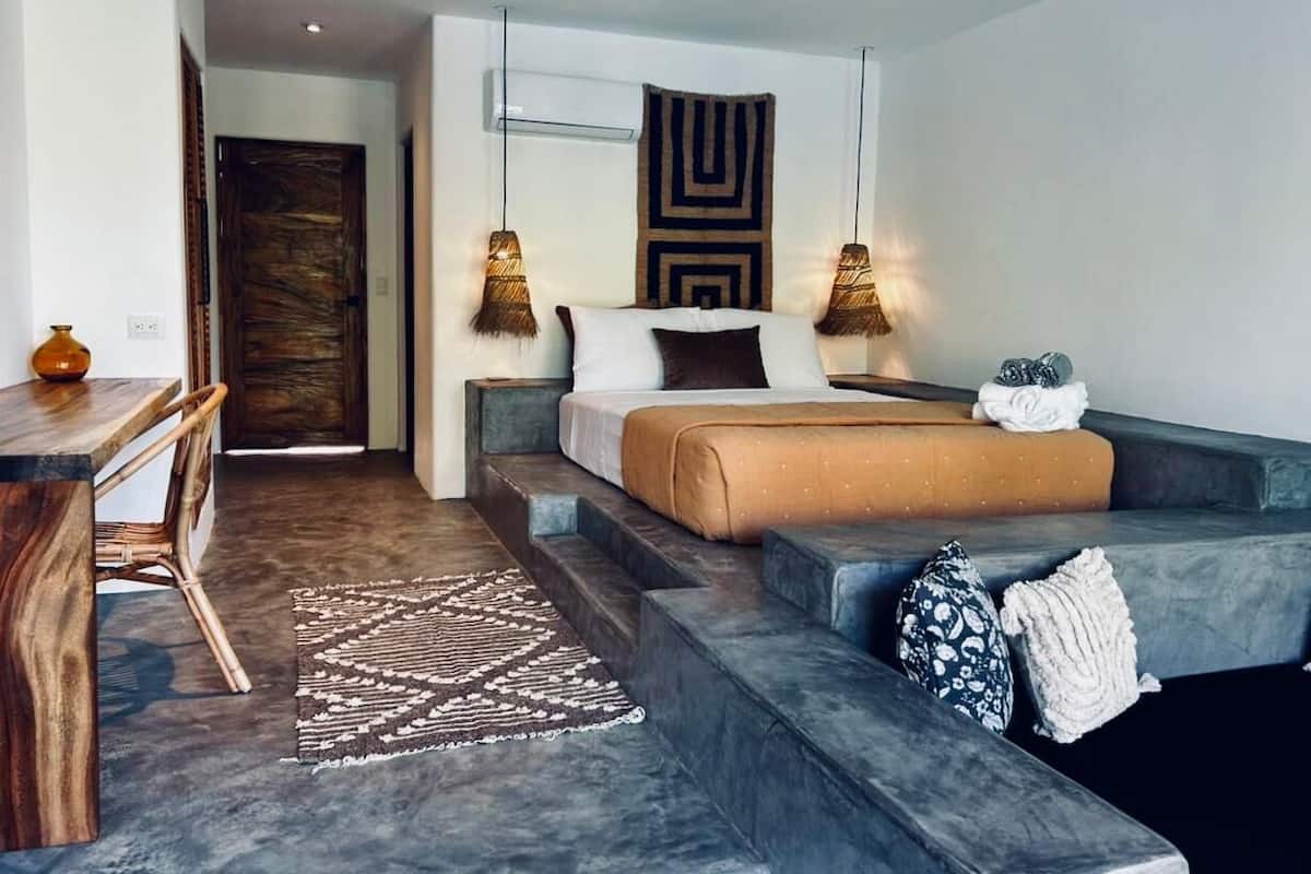 A modern bedroom with a bed, a desk, and a chair. There is a rug on the floor and a wall hanging above the bed.
