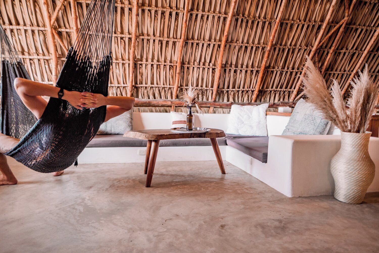 Una habitación con techo de bambú trenzado, una hamaca, un sofá, una mesa de centro y un florero con plantas secas.