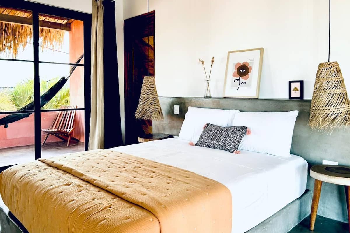 A large bed with a beige blanket, white pillows, and a grey headboard. There are two wall-mounted lamps and a framed picture above the bed.