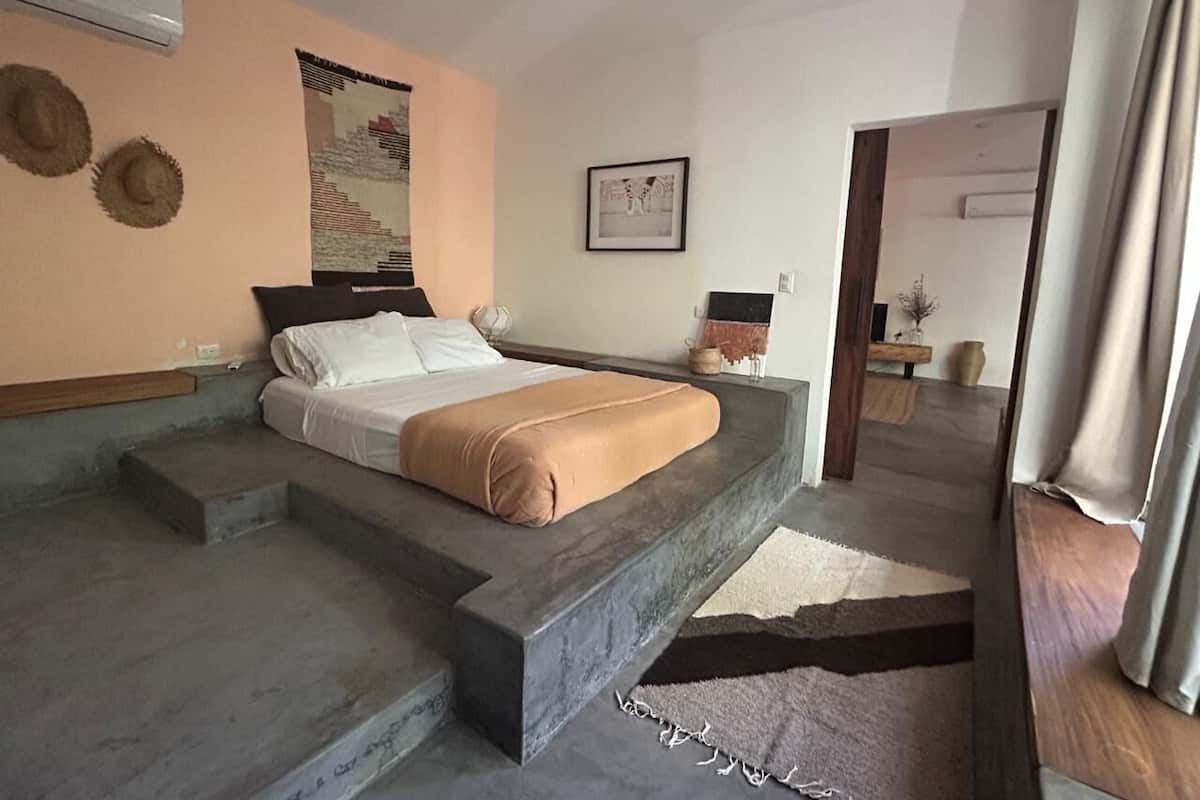 A bedroom with a bed, a headboard, a nightstand, a wall-mounted air conditioner, a framed picture, and a doorway leading to another room.