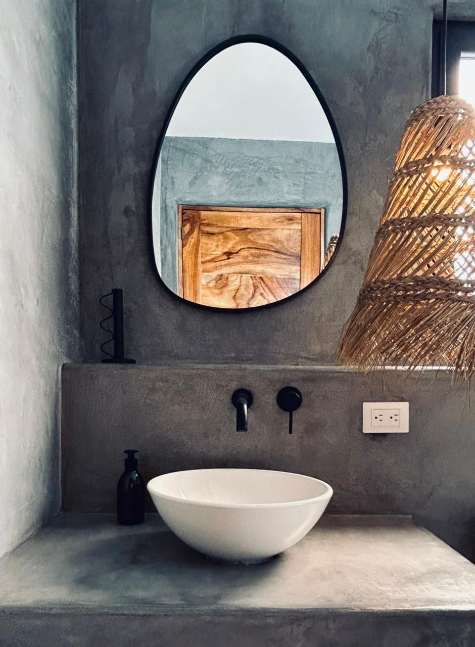 Un baño con un espejo ovalado grande, un panel de madera, un lavamanos blanco y una lámpara de fibra trenzada.