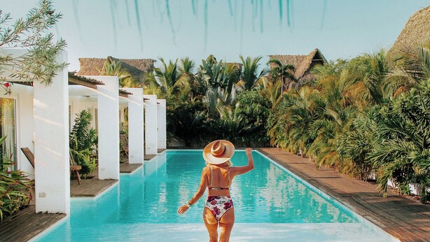 A woman walking away from a pool with a tropical setting, including palm trees and a wooden deck.
