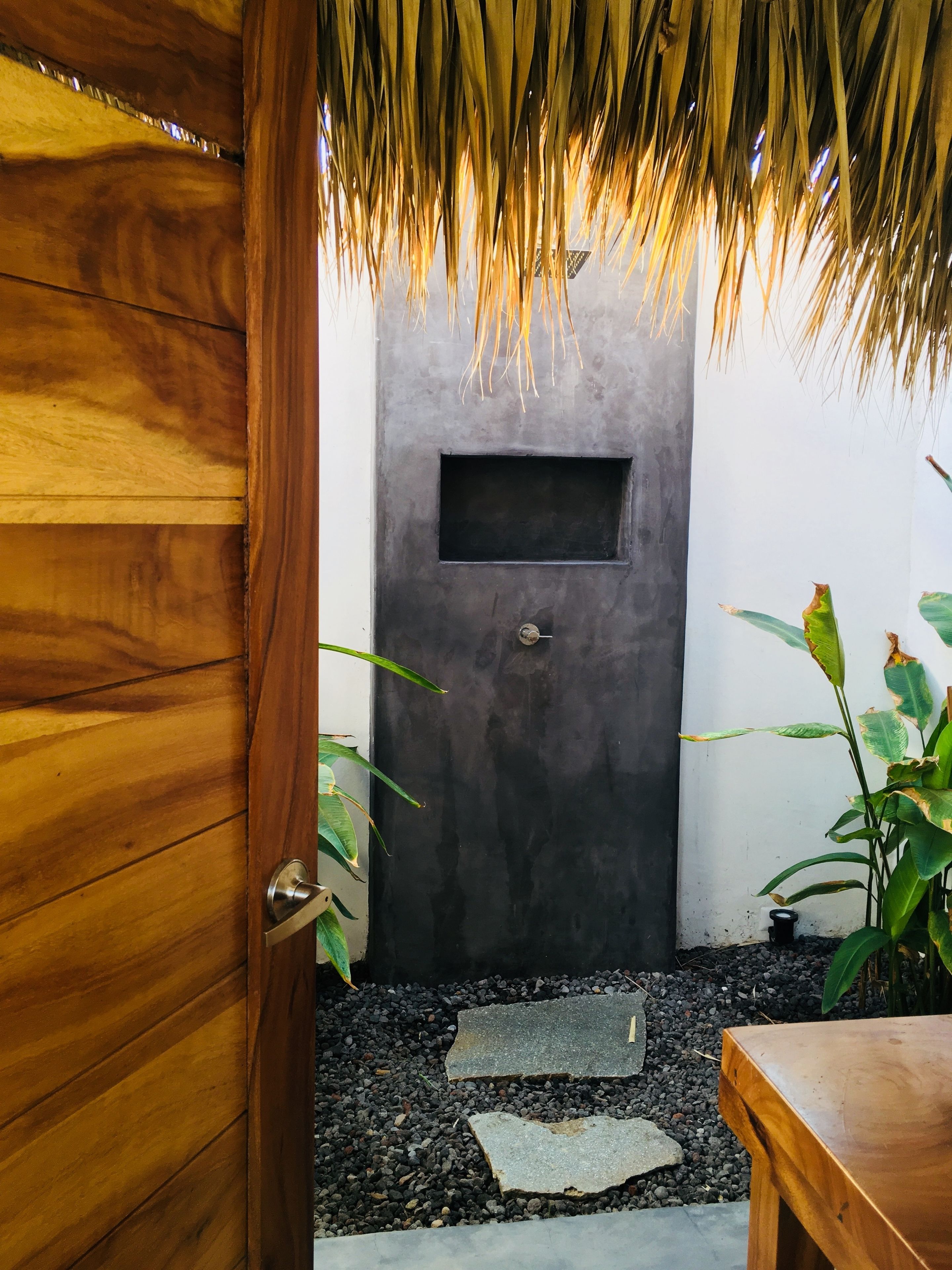 A wooden door leading to a room with a dark gray wall, a small window, and a thatched roof. There is a stone pathway and a wooden bench outside.