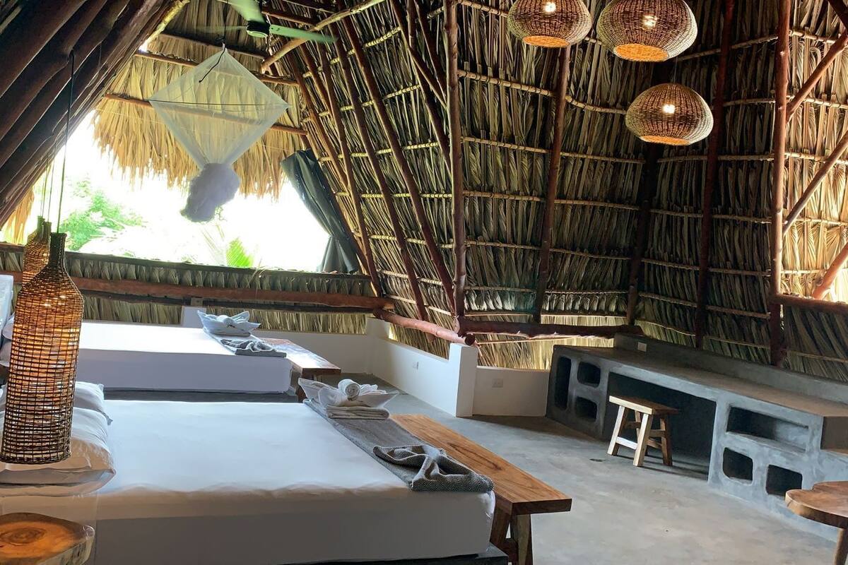 A room with a thatched roof, two beds, a wooden bench, and woven light fixtures.