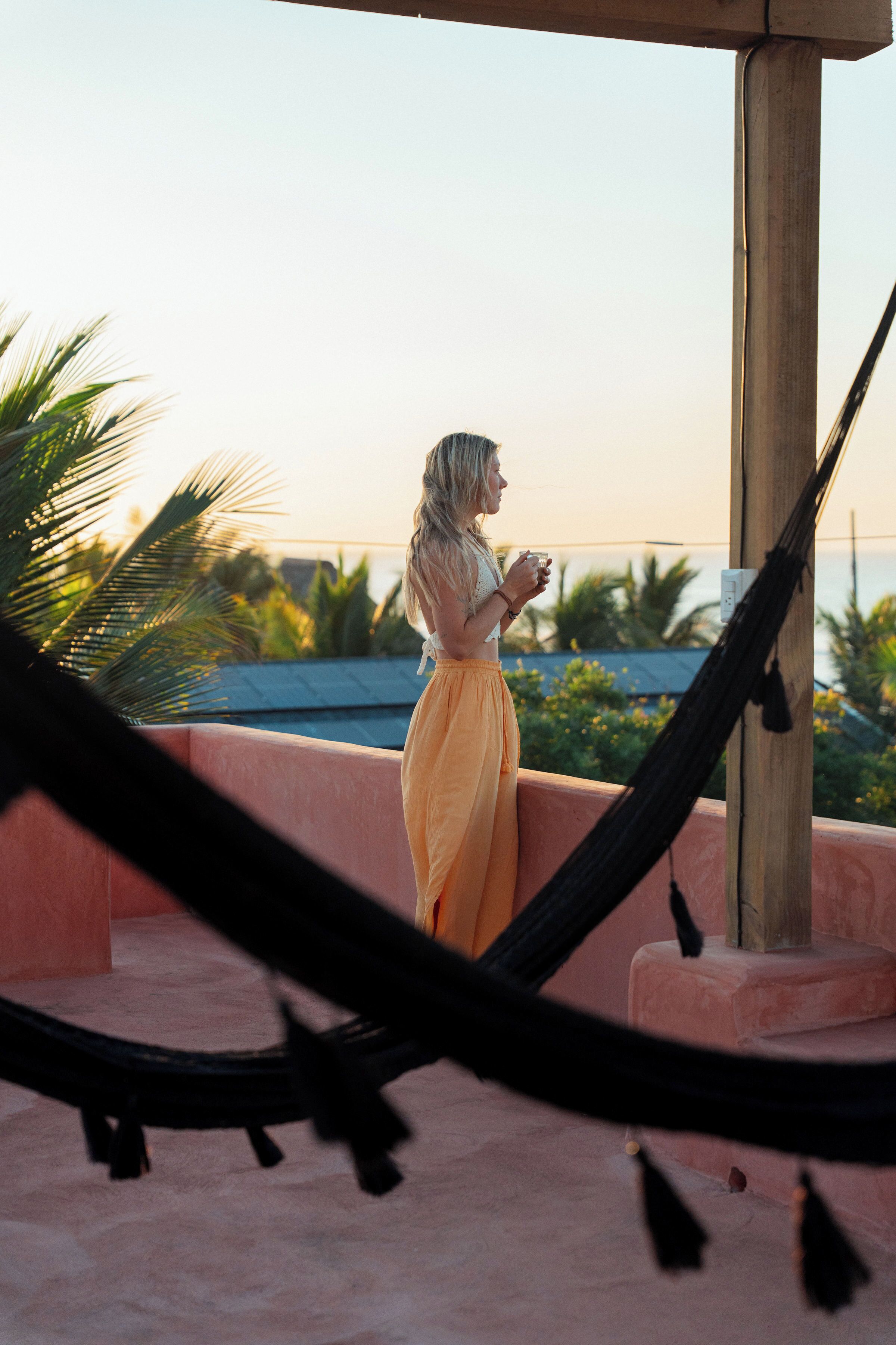 A person standing on a balcony with a hammock, overlooking a coastal area with palm trees and a clear sky.