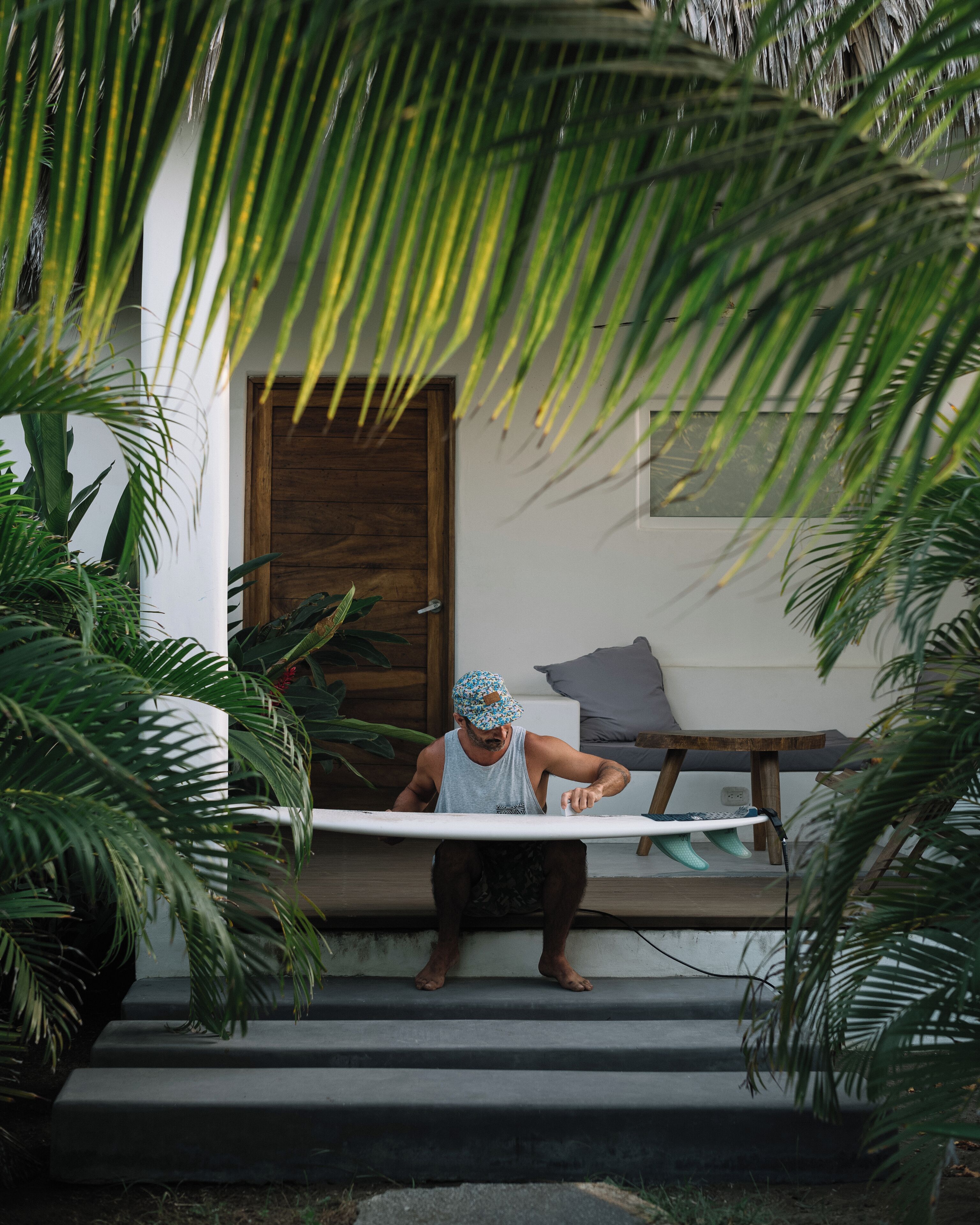 A person sitting on outdoor steps with a surfboard, surrounded by lush greenery and a wooden door.