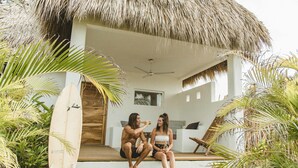 A couple sitting on a porch with a thatched roof, surrounded by tropical plants.